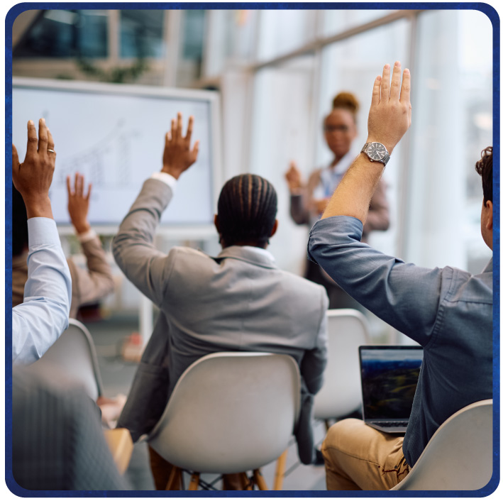 Audience members raise hands to engage with a presenter in a modern classroom setting.