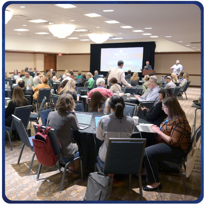 Attendees seated in roundtable groups during a large conference session with presentation at front. 