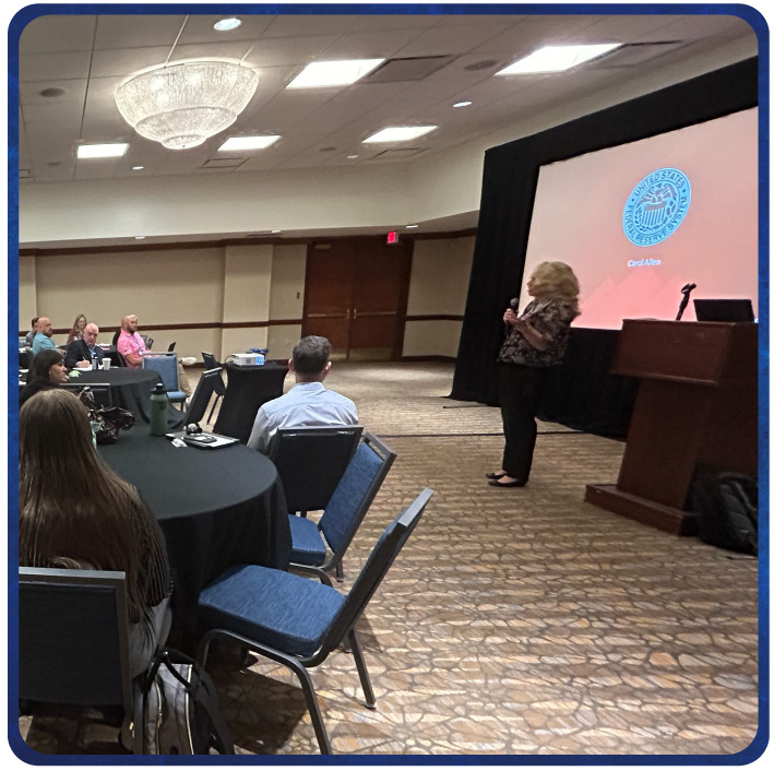 Presenter speaks to attendees seated at round tables, displaying slides on a screen behind presenter 