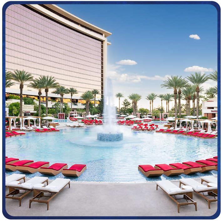 Resort pool with central fountain, palm trees, and red lounge chairs in front of a high-rise hotel. 