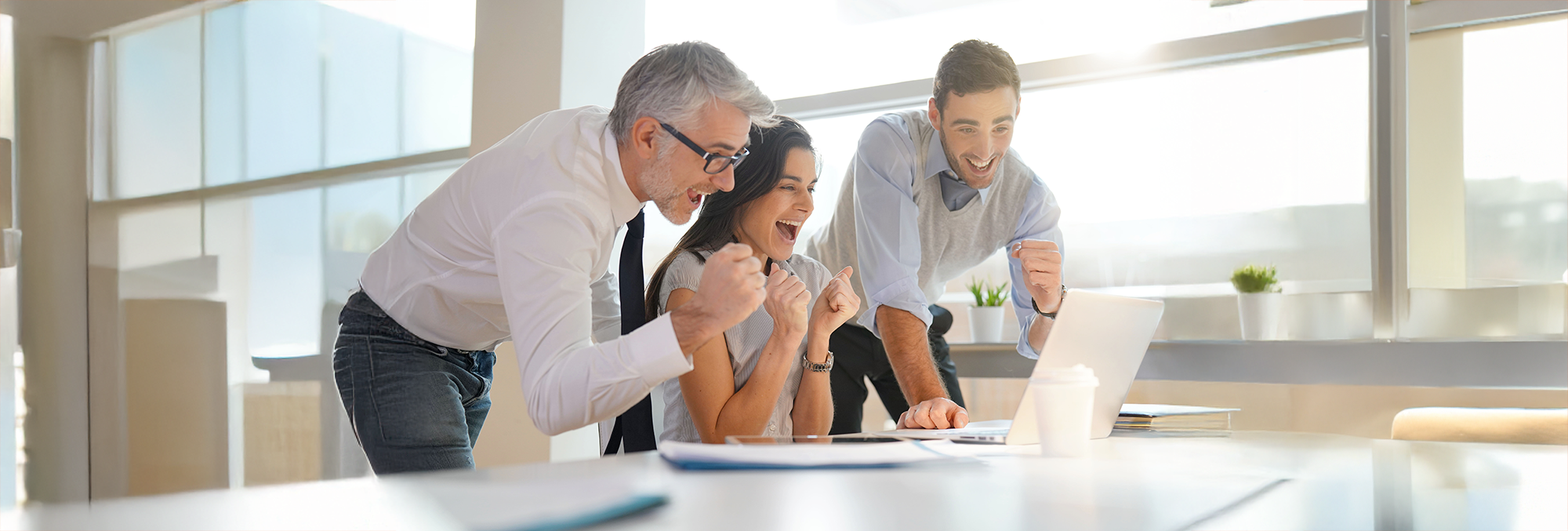 Three colleagues celebrate success while looking at a laptop together in a bright office.