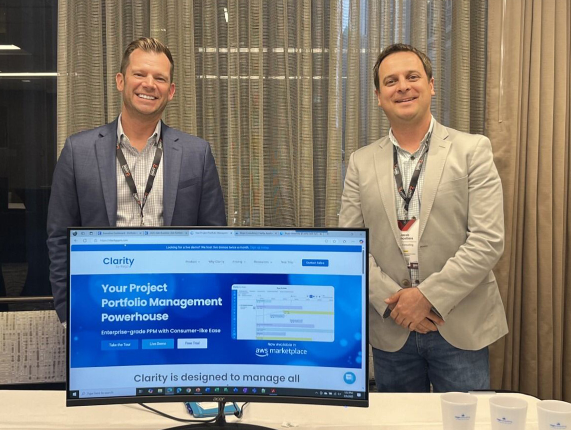 Two men stand smiling behind a monitor displaying Clarity project portfolio management software. 