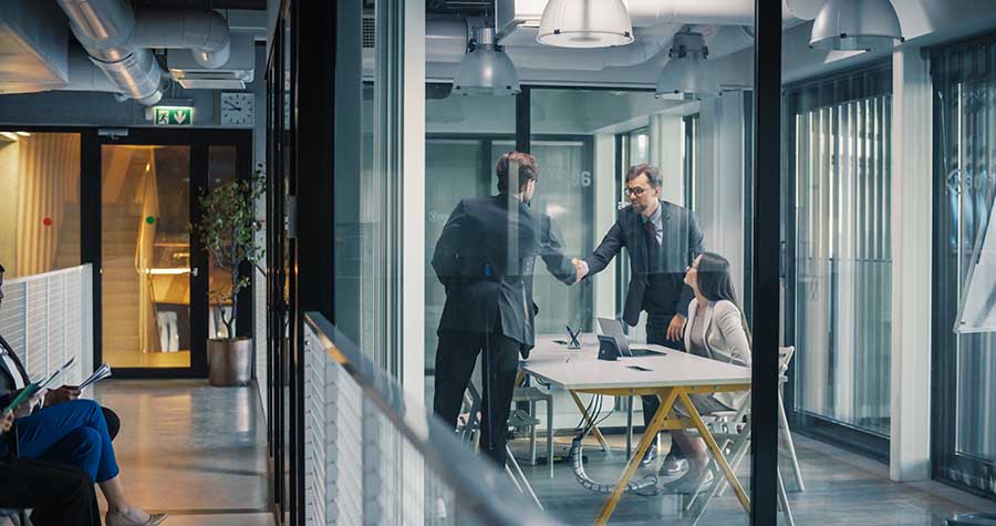Two colleagues shake hands during a meeting with a third colleague in a glass-walled office.